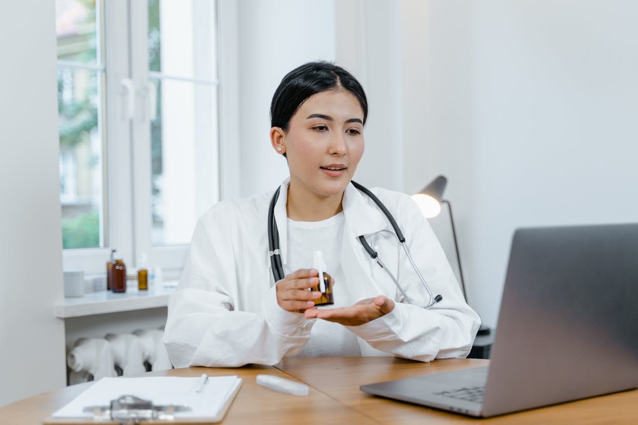 A young female doctor in a white coat on a video call, discussing medication with a patient.